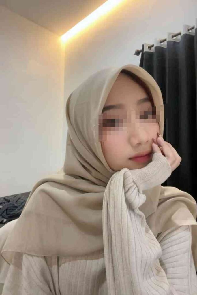 KL Escort Girl Young Muslim woman wearing a beige hijab and sweater, resting her chin on her hand. Sex Escort Malaysia Girl Service